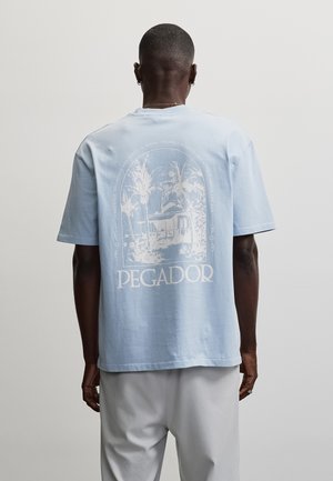 Bărbat purtând un tricou bleu deschis cu un grafic alb al unui resort tropical și cu cuvântul „PEGADOR” pe partea din spate, asortat cu pantaloni deschiși la culoare.