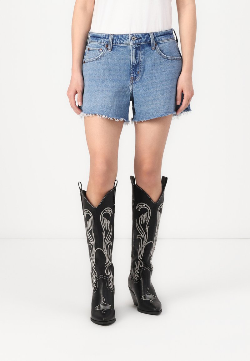 Zwarte leren cowboylaarzen met geborduurde witte details, hoge schacht en spitse neuzen, gecombineerd met versleten spijkerbroekshorts.