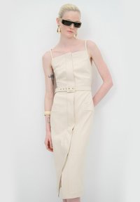 Robe beige ajustée avec bretelles spaghetti, taille ceinturée et fente avant. Matériau texturé avec des coutures décoratives et deux poches latérales.