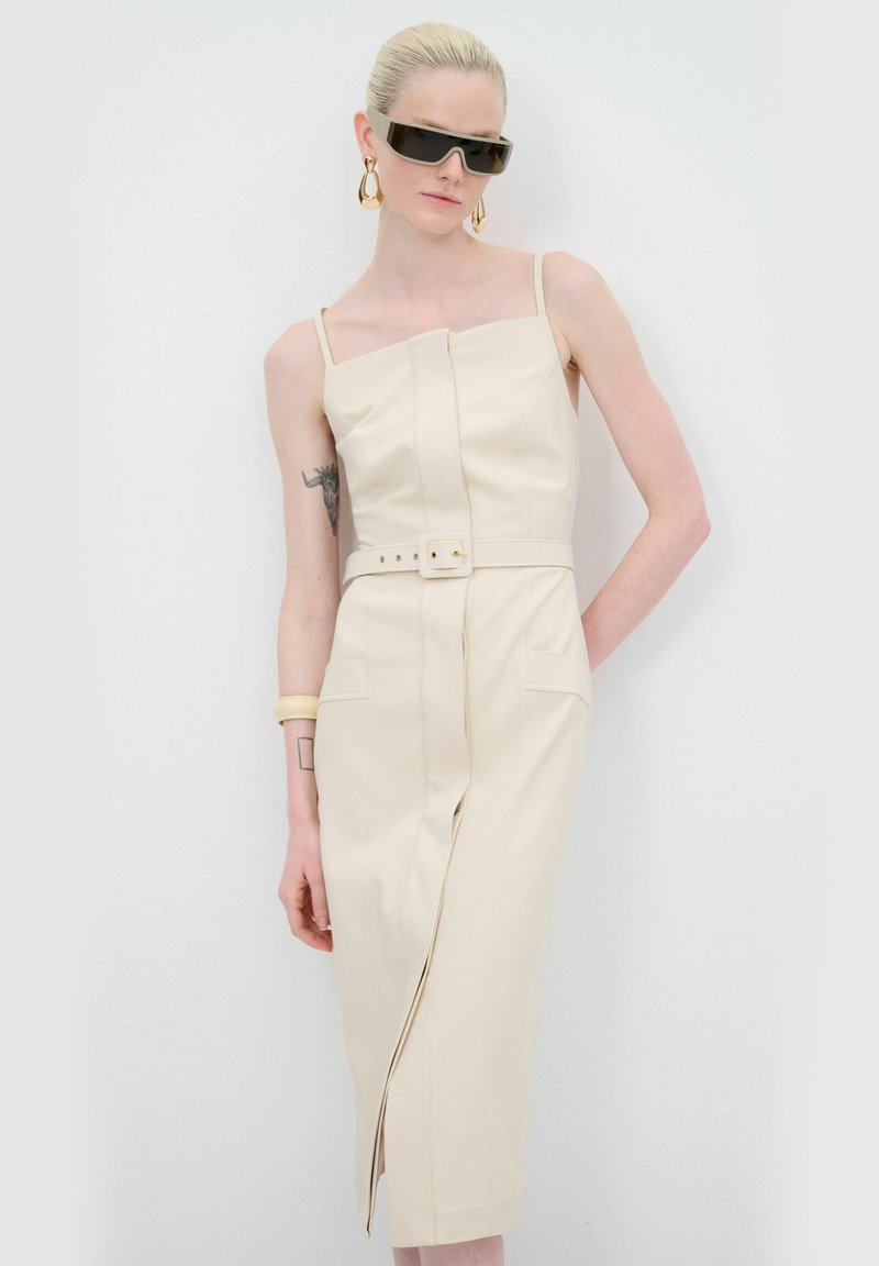 Robe beige ajustée avec bretelles spaghetti, taille ceinturée et fente avant. Matériau texturé avec des coutures décoratives et deux poches latérales.