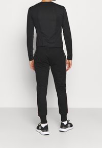 Schwarzes langärmliges Oberteil und Hose aus einem glatten, dehnbaren Stoff. Die Hose hat seitliche Streifen und gerippte Bündchen. Sportliche Schuhe runden das Outfit ab.