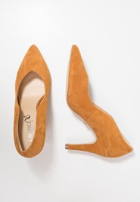 Světlé nubukové lodičky na vysokém podpatku s špičatou špičkou, výřezy po stranách a úzkým podpatkem. Interiér má béžovou stélku s logem.