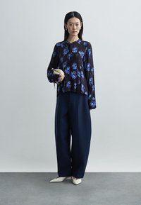 Fließende schwarze Bluse mit blauem Blumenmuster, kombiniert mit weiten marineblauen Hosen. Das Modell hält eine kleine beigen Clutch und trägt spitze weiße Schuhe.