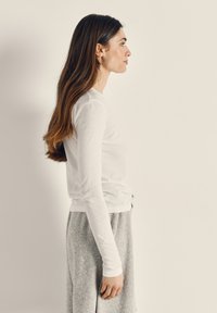 Pitkät ruskeat hiukset omaava nainen, joka seisoo sivuprofiilissa valkoisessa pitkähihaisessa paidassa ja harmaissa housuissa yksinkertaisen vaalean taustan edessä.