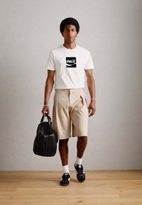 Witte t-shirt met zwart Coca-Cola logo, beige geplooide shorts, zwarte tas, zwarte sneakers met witte accenten, witte sokken. Achtergrond van houten vloer.