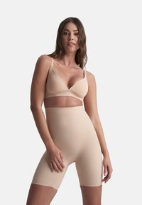 Donna che indossa shorts modellanti a vita alta color nude e un reggiseno abbinato, in piedi contro uno sfondo bianco semplice con una postura rilassata.