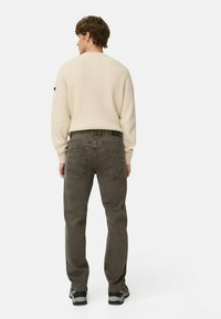 Man in een beige geribbelde trui, olijfgroene jeans en zwarte sneakers, staand met zijn rug naar de camera tegen een effen witte achtergrond.