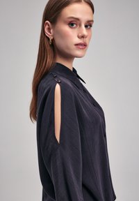 Ciemnoniebieska bluzka z guziczkami na ramionach i wyciętymi rękawami. Miękka, teksturowana tkanina oraz klasyczny kołnierz. Luźny krój.