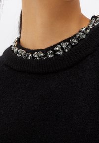 Zwarte trui met een gestructureerde afwerking, voorzien van een kraag die is versierd met sprankelende, veelvlakkige strass-steentjes in verschillende maten.
