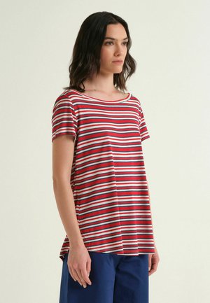 Femme aux cheveux foncés portant un t-shirt à manches courtes à rayures horizontales rouges, blanches et bleues et un pantalon bleu foncé, debout devant un fond clair.