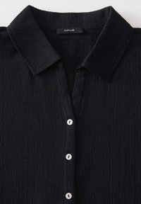 Czarna teksturowana koszula z kołnierzykiem i trzema białymi guzikami z przodu, z widoczną metką marki OPUS wewnątrz kołnierza.