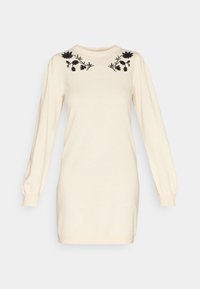 Robe tricotée crème avec des manches longues, un col rond et un motif floral noir brodé sur les épaules. Texture douce, coupe décontractée.