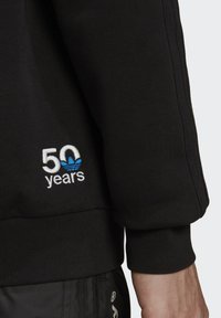 Sort sweatshirt med ribbede manchetter, der viser hvid og blå broderet tekst "50 år" med et adidas-logo over. Blød stoftekstur.