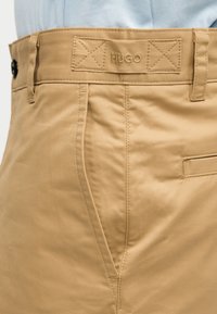 Κοντινή λήψη μπεζ χακί παντελονιού με πλαϊνή τσέπη και ραμμένη ετικέτα «HUGO» στη μέση, φορεμένο με ανοιχτό μπλε πουκάμισο.