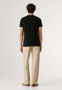 Homme aux cheveux bouclés, debout de dos, portant un t-shirt noir, un pantalon beige et des chaussures vertes et blanches sur un fond uni.