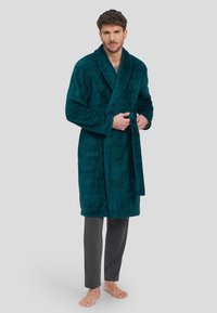 Bata de baño de felpa en color verde azulado con patrón texturizado, diseño envolvente y cinturón, combinada con pantalones de pijama grises. El modelo está descalzo, de pie sobre un fondo gris.