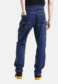 Pantalons de travail en denim bleu foncé, comportant plusieurs poches, des coutures contrastées et une taille ajustée. Finition en tissu durable avec texture.