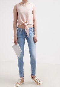 Top corto con textura en rosa claro y lazo en la parte delantera, combinado con jeans ajustados azules claros. La modelo lleva zapatillas metálicas doradas y sostiene un bolso gris.