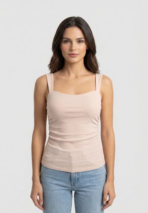 Jeune femme aux longs cheveux foncés portant un haut sans manches côtelé rose clair et un jean bleu clair, sur un fond uni.