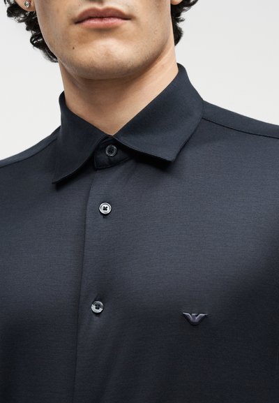 Κοντινό πλάνο ενός ατόμου που φοράει σκούρο πουκάμισο με κουμπιά και μικρό κεντημένο λογότυπο στο στήθος και σκουλαρίκια καρφάκια.