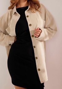 Beige Kordhemdjacke mit Knöpfen an der Vorderseite und einer Schultertasche, kombiniert mit einem taillierten schwarzen Rippenkleid. Sanfte Texturen.