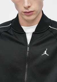 Μαύρο αθλητικό τζάκετ με φερμουάρ και ριμπ κολάρο, με άσπρες λεπτομέρειες και λογότυπο Jumpman στο αριστερό στήθος. Λεπτό ύφασμα, εφαρμοστό σχέδιο.