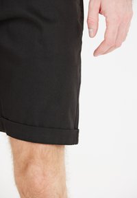 Svarta bomullsshorts med vikt kant, slät textur och en nära passform, visade tillsammans med en hand som vilar på tyget.