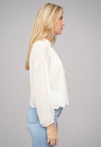 Hvid bluse med gennemsigtige, svævende ærmer og ståkrave, parret med lyseblå tætsiddende jeans. Enkel og elegant design.