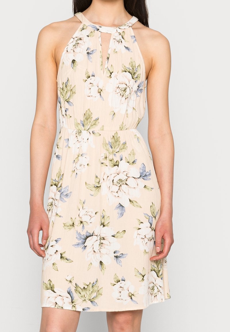 Robe beige clair avec un motif floral en blanc et bleu, dotée d'un col halter, d'un détail en forme de clef et d'une taille cintrée. Tissu léger.