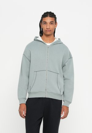Jonge man met een gevlochten kapsel, gekleed in een lichtgrijze zip-up hoodie en zwarte broek, tegen een eenvoudige witte achtergrond.