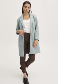 Ljusblå lång kappa av mjukt tyg med slagkrage, kombinerad med bruna smala byxor och svarta loafers. Vit spets-topp under.
