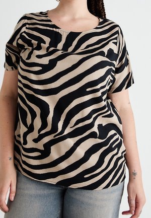 Kobieta ubrana w beżową bluzkę z krótkim rękawem z czarnymi abstrakcyjnymi paskami zebry oraz jasnoniebieskie dżinsy, stojąca na jednolitym białym tle.