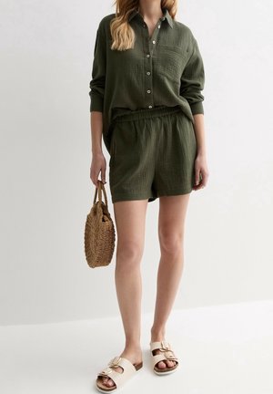 Femme portant une chemise ample vert olive boutonnée et un short, tenant un sac à main rond tissé, et portant des sandales beiges à boucle sur fond blanc.