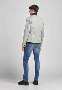 Jachetă din nubuc gri deschis cu guler înalt, detaliu de yoke pe spate, asortată cu blugi slim-fit de un albastru închis și pantofi negri cu șireturi.