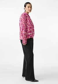 Pink and red patterned long-sleeve top with button closures, paired with black wide-leg trousers and glossy black heels. Smooth texture.