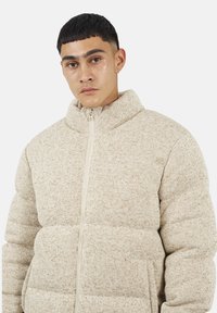 Giacca imbottita beige con colletto alto, chiusura con zip, e tessuto testurizzato. Presenta un design a pannelli cuciti e tasche laterali.