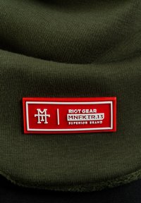Olive green knit fabric with a prominent red rubber label featuring white text: "RIOT GEAR MNFKTR.13 SUPERIOR BRAND." Soft texture.
