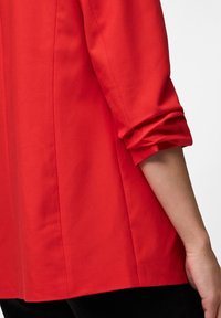 Chemise rouge avec un revers plié et une texture lisse, de coupe droite, mettant en valeur des lignes épurées et des détails de couture minimalistes.