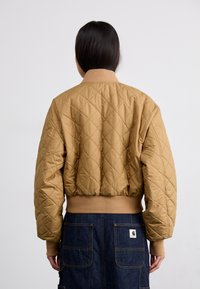 Chaqueta bomber acolchada en marrón claro con cuello y puños de canalé, con una silueta corta. Llevada sobre una falda de mezclilla oscura con bolsillos.
