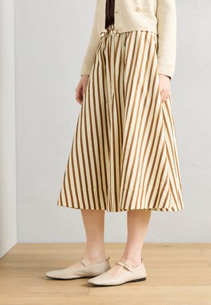 Kvinde iført beige og brunt lodret striped midi-nederdel, cremefarvet cardigan og beige flade Mary Jane-sko, stående på trægulv.