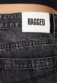 Šedé džíny z denimu s opotřebovanou texturou. Obsahují bílou visačku s nápisem "RAGGED" tučným černým písmem. Viditelné detaily šití.