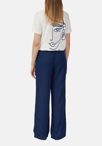 Weißes Kurzarm-T-Shirt mit einer schwarzen Lin Zeichnung eines Gesichts auf der Rückseite, kombiniert mit navyblauen weiten Hosen.
