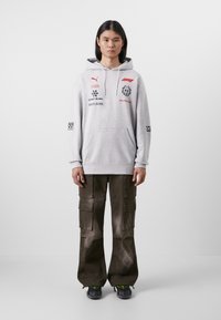Hanorac gri cu glugă, cu logo-uri, buzunar frontal și șnururi albe, asortat cu pantaloni cargo închisi la culoare, cu multiple buzunare și fermoare.