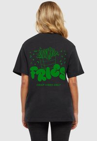 Must särkjuhendustega must T-särk, millel on roheline graafika friikartulite konteinerist, all tekst "FRIES" ja "CRISP VIBES ONLY".