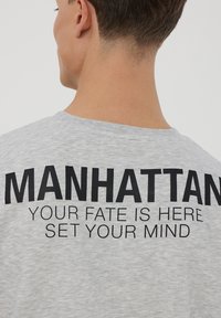 Maglietta in cotone grigia con grande testo nero che riporta la scritta "MANHATTAN" e un testo più piccolo sotto. Design semplice con colletto a girocollo, maniche corte, stile casual.