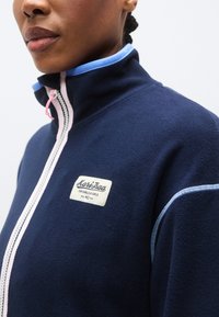 Το μπουφάν από φλίς σε ναυτικό μπλε διαθέτει ψηλό γιακά, ροζ φερμουάρ, ανοιχτό μπλε λεπτομέρειες και ένα διακοσμητικό έμβλημα με κείμενο. Μαλακή υφή και αθλητικός σχεδιασμός.