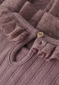 Jasnoliliowy top z teksturowanej tkaniny z pionowymi paskami, koronkowymi falbanami wzdłuż dekoltu i małym drewnianym guzikiem jako akcentem.