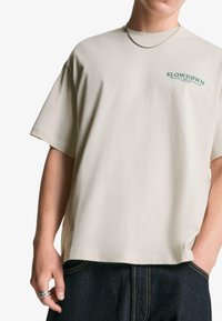 Persoană purtând un tricou oversized bej cu inscripția "SLOWDOWN" pe piept și jeans închisi la culoare, accesorizată cu un colier din argint și un inel.