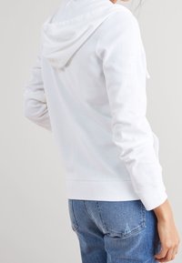 Felpa bianca con cappuccio, con texture a coste, maniche lunghe e tasca a marsupio. Abbinata a jeans in denim chiaro strappati.
