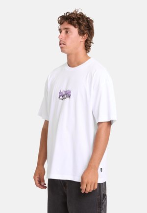 Junger Mann mit lockigem Haar, der ein weißes übergroßes T-Shirt mit lila grafischem Text und schwarze Hose trägt, nach links auf einem einfarbigen Hintergrund blickend.